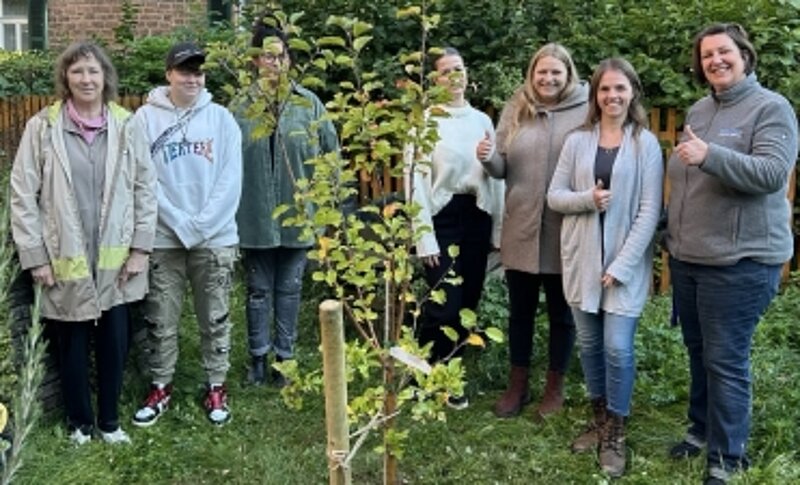 Eine Menschengruppe steht hinter einem neu geplanzten Apfelbaum.
