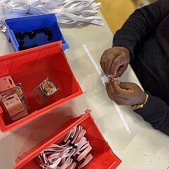 Bild aus dem Arbeitsbereich Montage. Man sieht Hände einer Person, die an einem Tisch Bänder und Ösen aus verschiedenen Kästen entnimmt und verbindet.