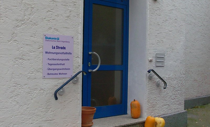 Eingangstür mit Schild: "La Strada, Wohnungsnotfallhilfe: Fachberatungsstelle, Tagesaufenthalt, Übergangswohnheim, Betreutes Wohnen"