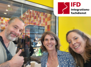 Drei Personen schauen freundlich lächelnd in die Kamera. Hinter ihnen ist ein Schaufenster und eine gelbe Wand zu sehen.